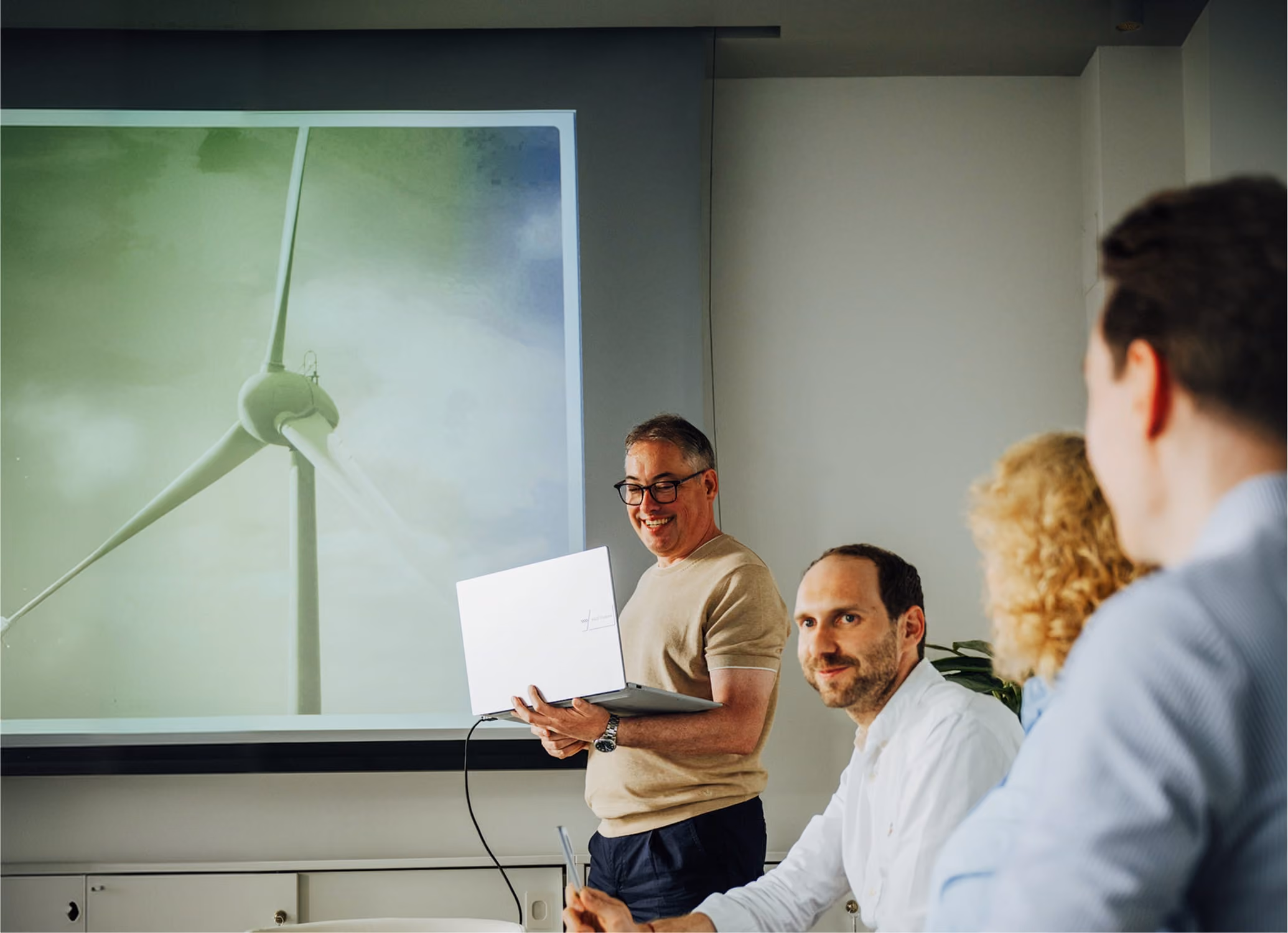 Mann präsentiert Windkraftanlage auf Projektor, drei Personen hören bei Meeting zu.